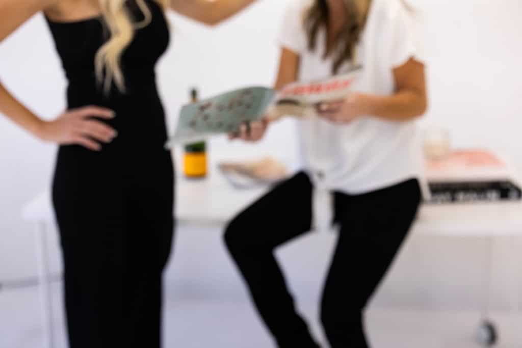 Blurred image of two women in conversation while holding an open magazine, with a bottle of champagne and stack of books in the background.
