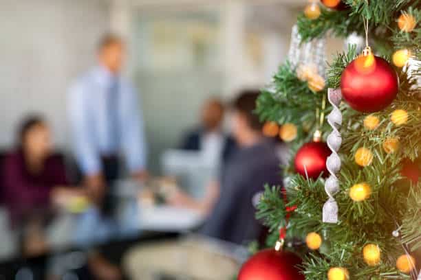 Close-up of a decorated Christmas tree with red and gold ornaments in the foreground, with a blurred office meeting happening in the background.